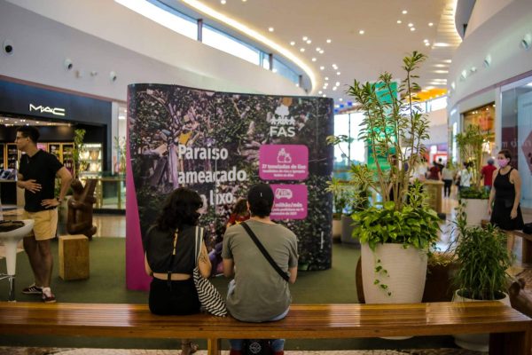 Pessoas sentadas em banco no shopping, vendo exposição realizada pela Fundação Amazônia Sustentável (FAS).